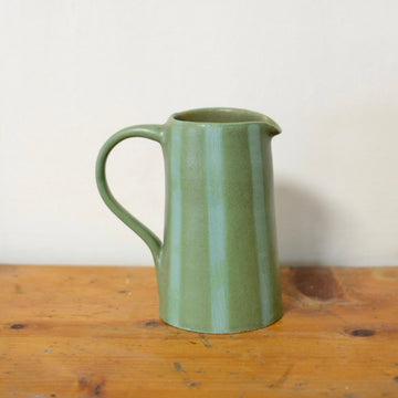 A green ceramic pitcher with a curved handle and spout, sitting on a rustic wooden table against a plain off-white wall.