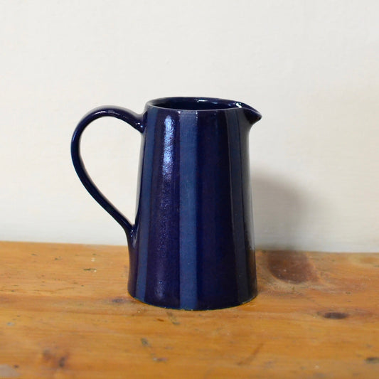 A dark navy blue striped glazed ceramic pitcher with a curved handle, resting on a rustic wooden surface against a plain off-white wall.