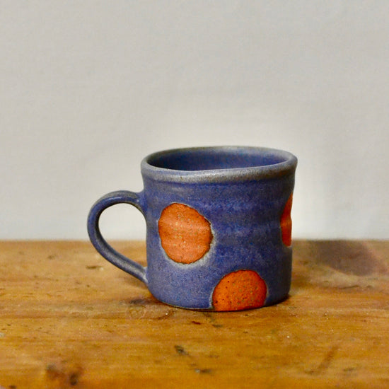 A small ceramic mug with a blue-gray glaze and coral polka dots, sitting on a wooden surface against a plain light grey wall.