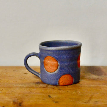 A small ceramic mug with a blue-gray glaze and coral polka dots, sitting on a wooden surface against a plain light grey wall.