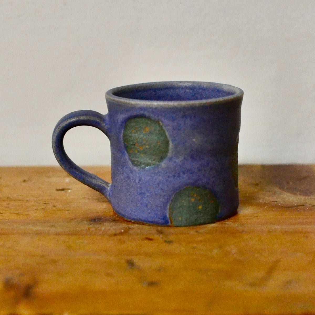 A handmade ceramic mug with a textured blue glaze and green polka dot accents, sitting on a rustic wooden surface against a plain light grey wall.