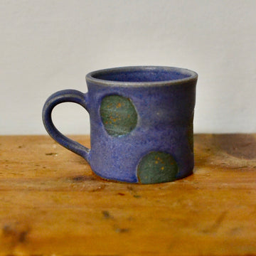 A handmade ceramic mug with a textured blue glaze and green polka dot accents, sitting on a rustic wooden surface against a plain light grey wall.