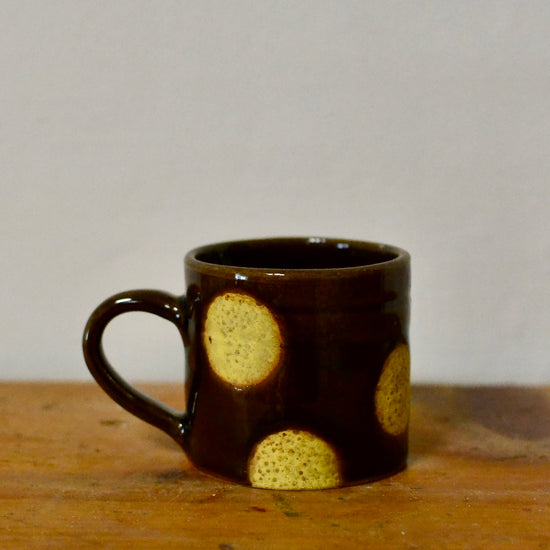A small dark brown ceramic mug with a handle, decorated with three textured yellow circular spots, sitting on a wooden surface against a plain light grey background.