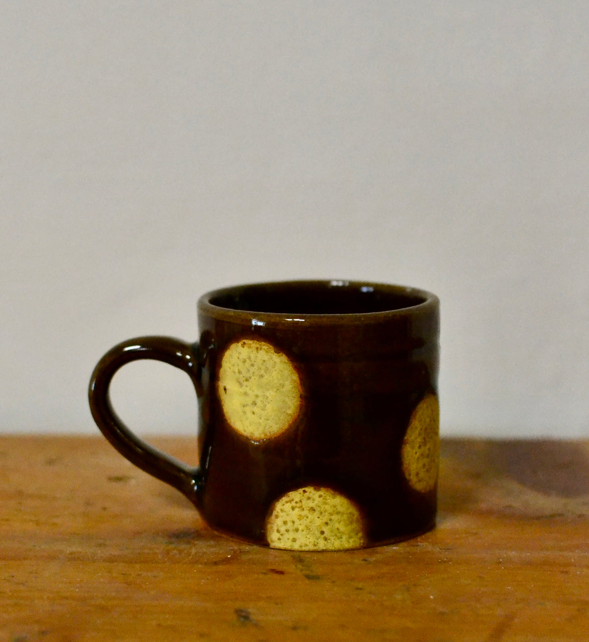 A small dark brown ceramic mug with a handle, decorated with three textured yellow circular spots, sitting on a wooden surface against a plain light grey background.