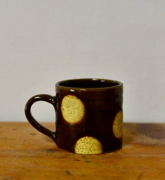 A small dark brown ceramic mug with a handle, decorated with three textured yellow circular spots, sitting on a wooden surface against a plain light grey background.