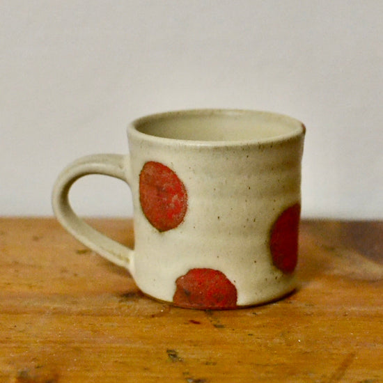 A handmade ceramic mug with a speckled beige glaze and large red polka dots, resting on a rustic wooden surface against a plain light gray wall.