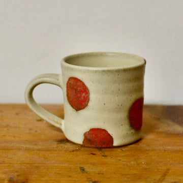 A handmade ceramic mug with a speckled beige glaze and large red polka dots, resting on a rustic wooden surface against a plain light gray wall.
