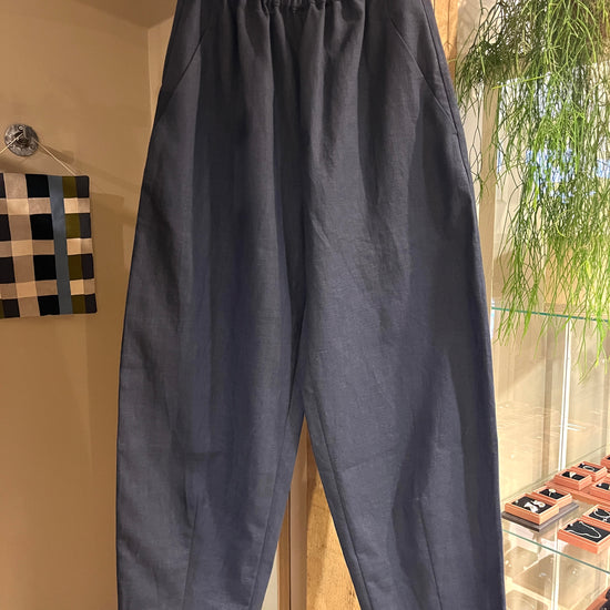 A pair of navy blue cropped trousers with an elastic waistband, hanging on a wooden hanger inside a boutique display.