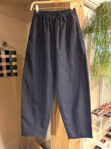 A pair of navy blue cropped trousers with an elastic waistband, hanging on a wooden hanger inside a boutique display.