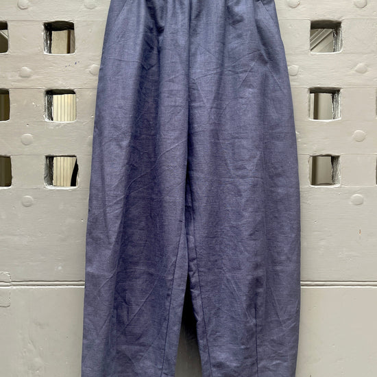A pair of loose, barrel leg trousers in a navy blue linen fabric, hanging from a light-colored wooden hanger against a textured grey metal door with square cutouts.