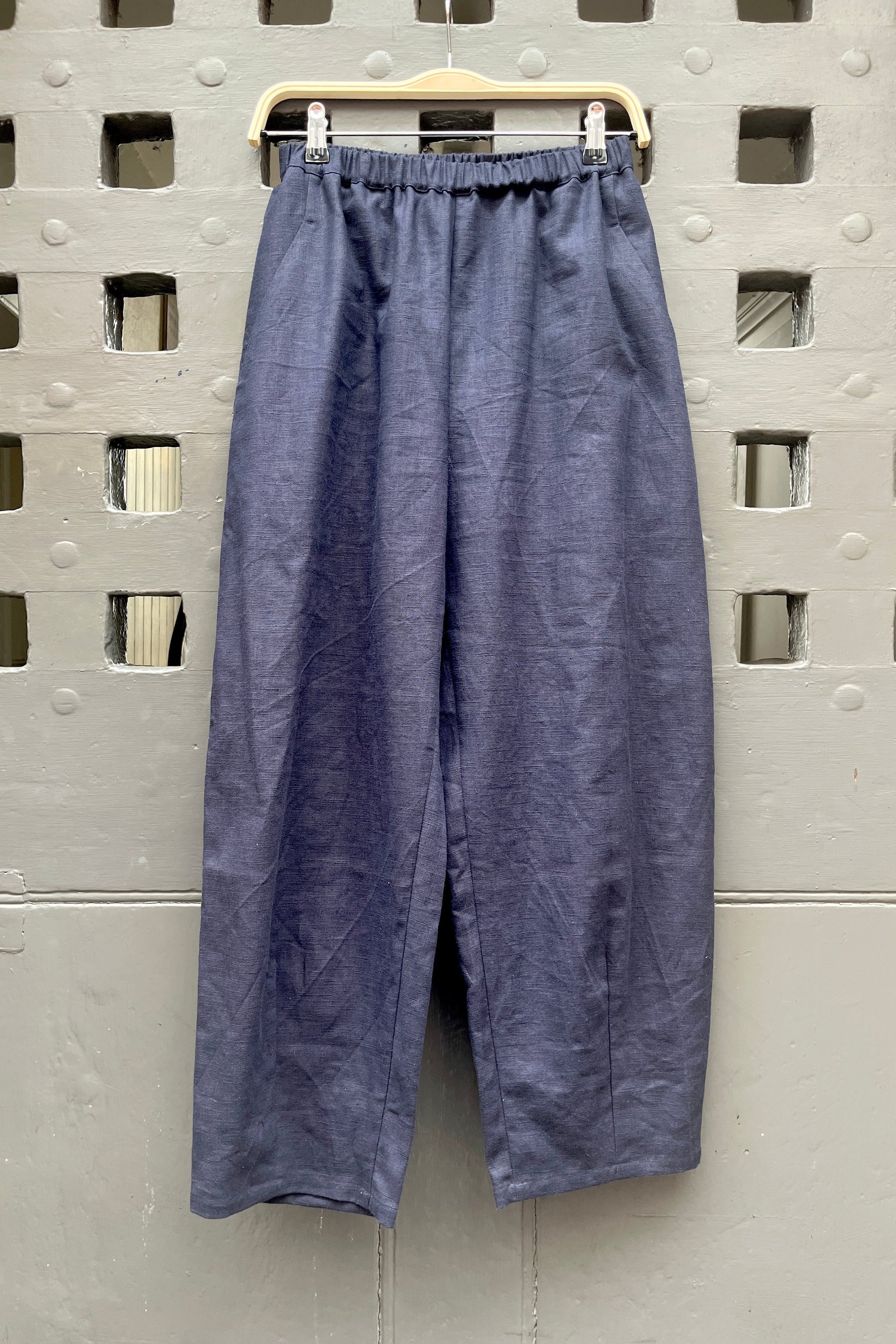 A pair of loose, barrel leg trousers in a navy blue linen fabric, hanging from a light-colored wooden hanger against a textured grey metal door with square cutouts.