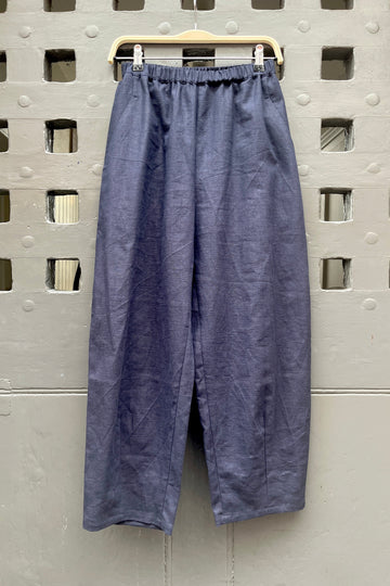 A pair of loose, barrel leg trousers in a navy blue linen fabric, hanging from a light-colored wooden hanger against a textured grey metal door with square cutouts.