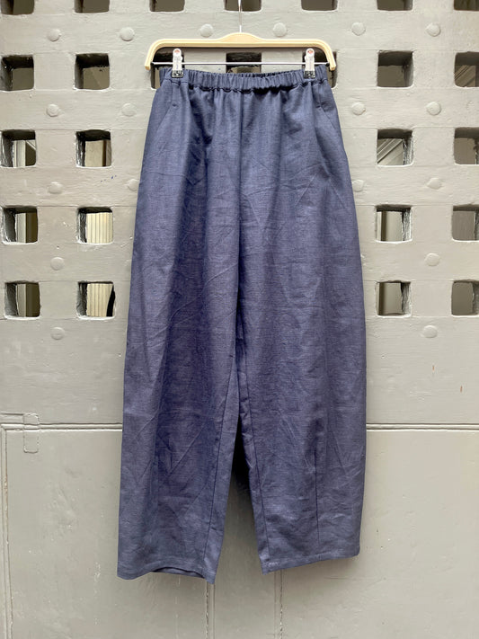 A pair of loose, barrel leg trousers in a navy blue linen fabric, hanging from a light-colored wooden hanger against a textured grey metal door with square cutouts.