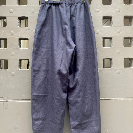 A pair of loose, barrel-leg trousers in a navy blue fabric, hanging from a light-colored wooden hanger against a textured grey door with a square-grid pattern.