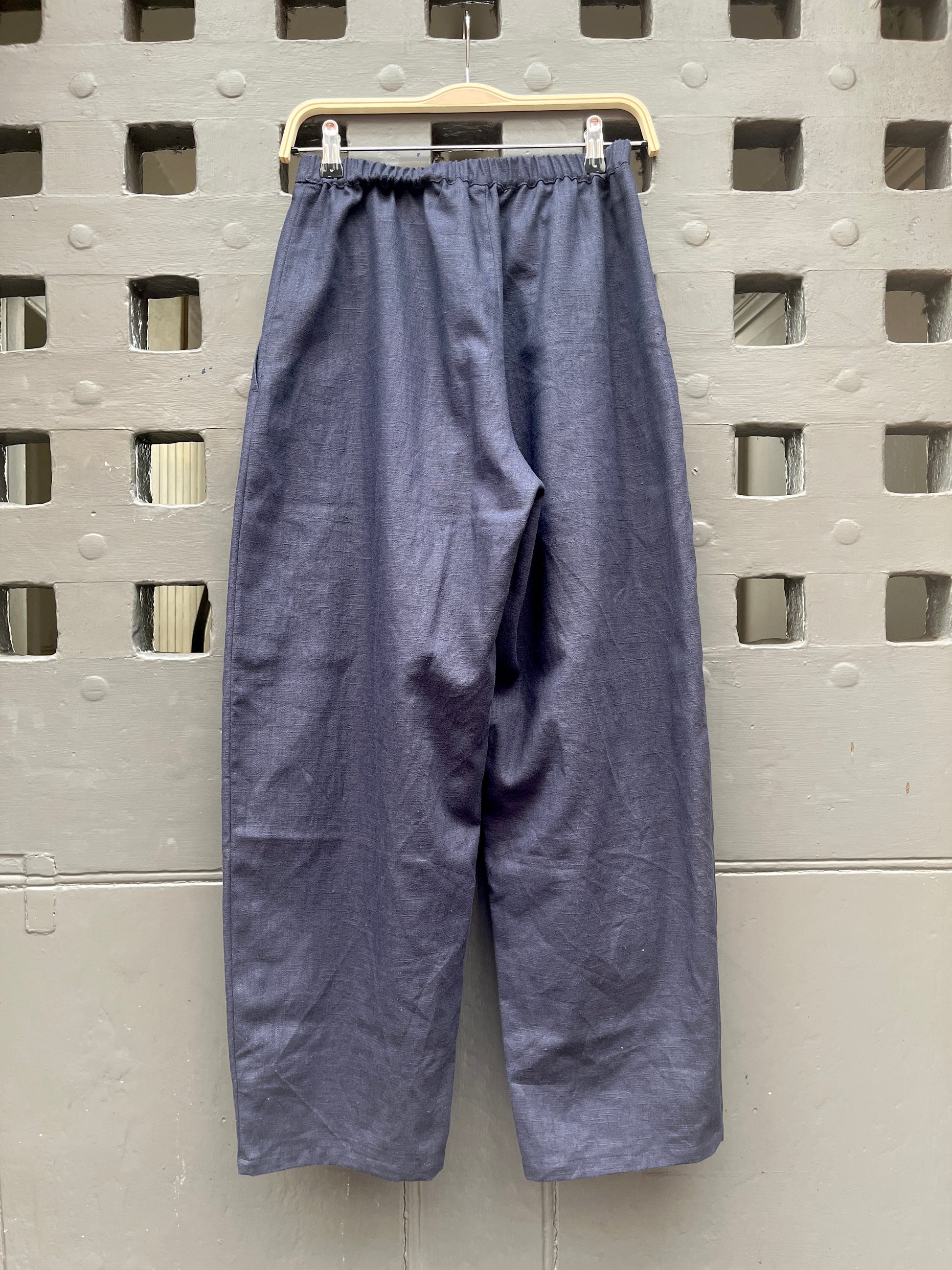 A pair of loose, barrel-leg trousers in a navy blue fabric, hanging from a light-colored wooden hanger against a textured grey door with a square-grid pattern.