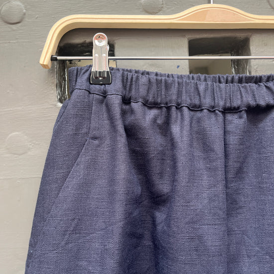Close-up of a pair of navy blue linen fabric trousers hanging on a wooden hanger against a grey door