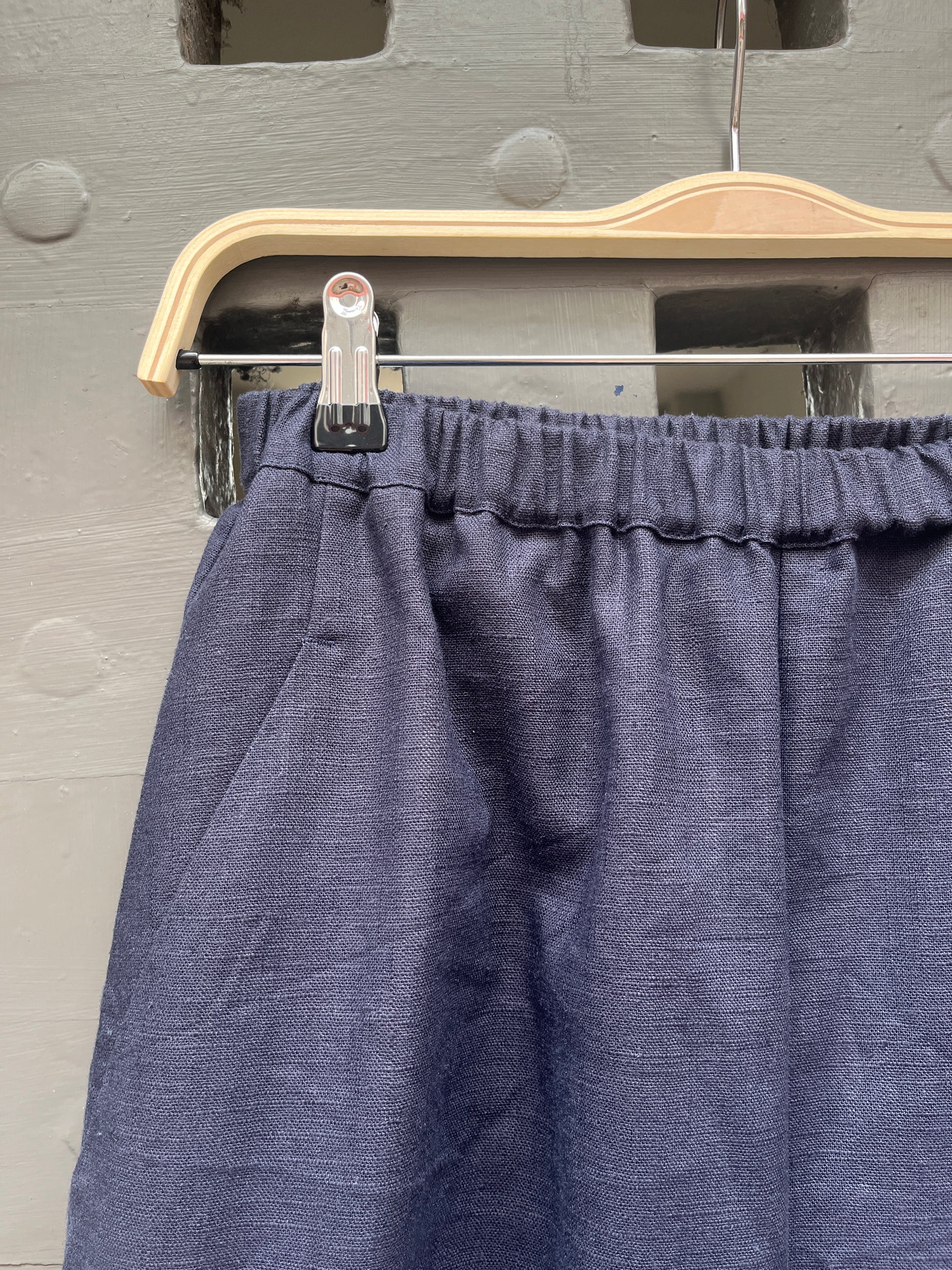 Close-up of a pair of navy blue linen fabric trousers hanging on a wooden hanger against a grey door