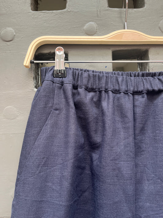 Close-up of a pair of navy blue linen fabric trousers hanging on a wooden hanger against a grey door