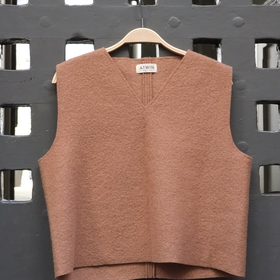 Camel coloured boiled wool sleeveless v-neck vest on a wooden hanger against a grey grid door