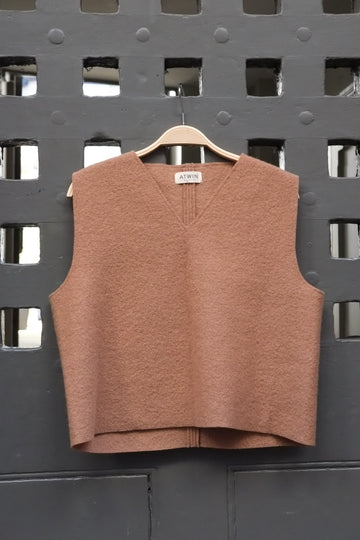 Camel coloured boiled wool sleeveless v-neck vest on a wooden hanger against a grey grid door