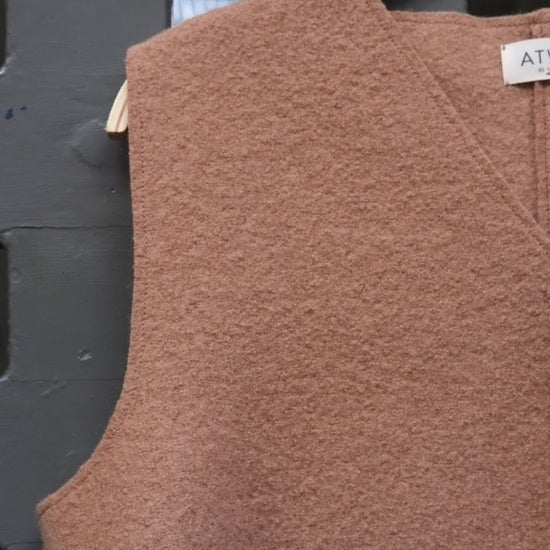 Camel coloured boiled wool sleeveless v-neck vest on a wooden hanger against a grey grid door