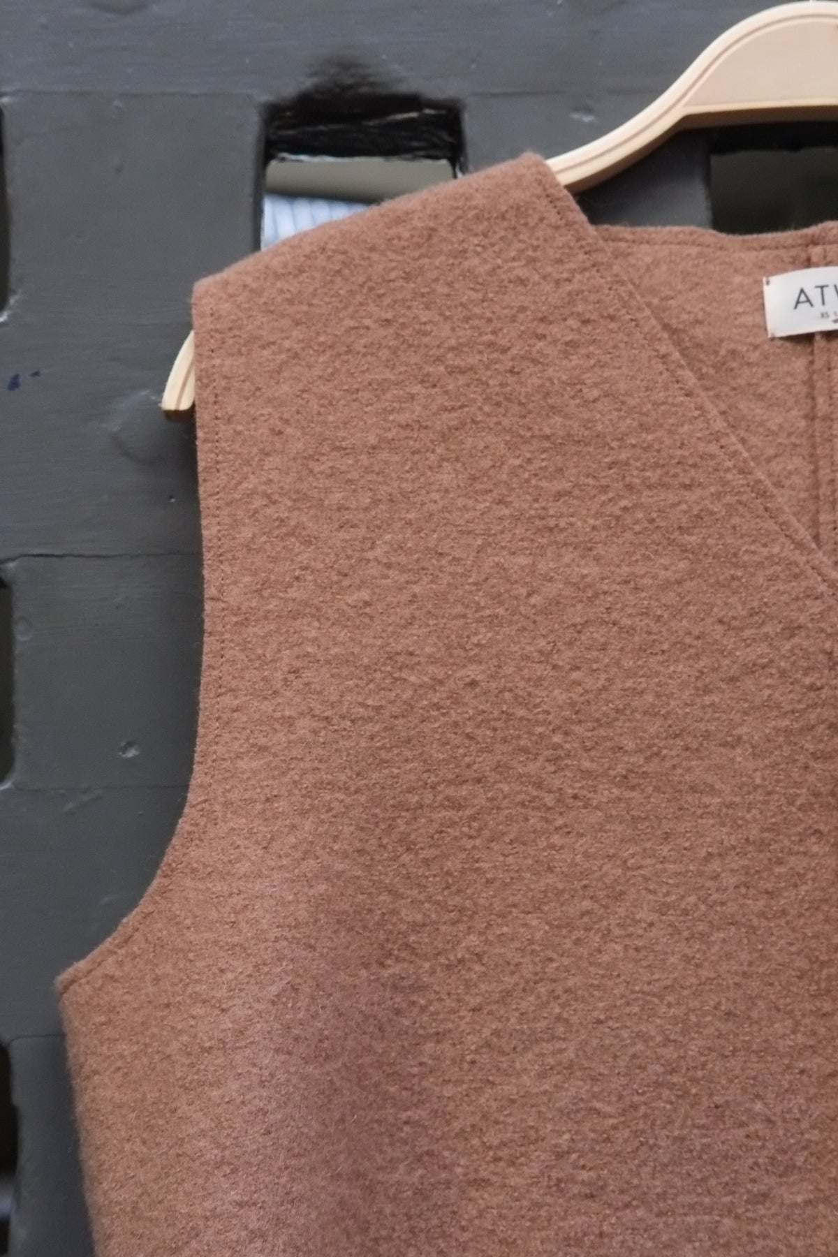 Camel coloured boiled wool sleeveless v-neck vest on a wooden hanger against a grey grid door