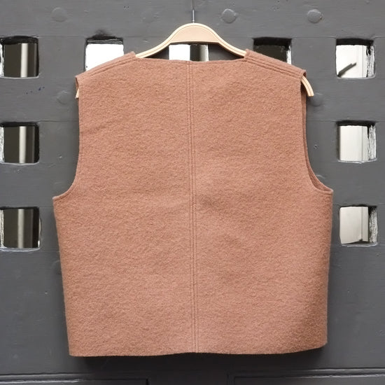Camel coloured boiled wool sleeveless v-neck vest on a wooden hanger against a grey grid door