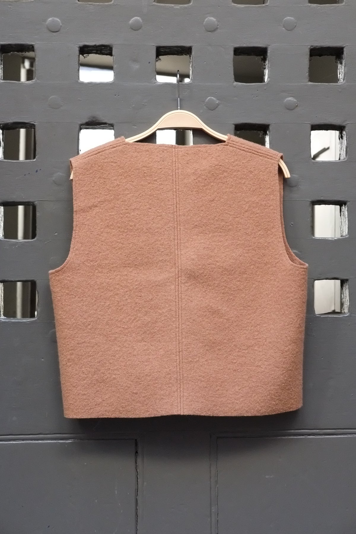 Camel coloured boiled wool sleeveless v-neck vest on a wooden hanger against a grey grid door
