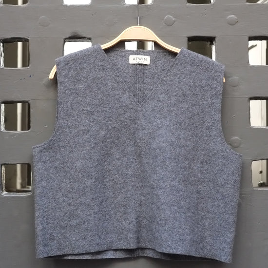 A sleeveless grey boiled wool vest hanging on a wooden hanger against a dark grey metal lattice door.
