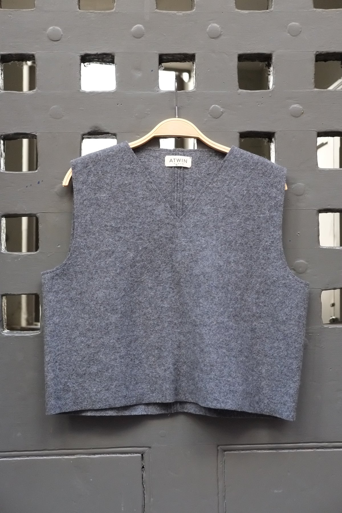 A sleeveless grey boiled wool vest hanging on a wooden hanger against a dark grey metal lattice door.