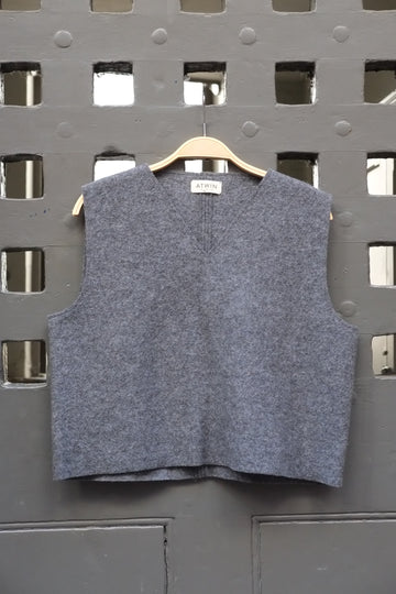 A sleeveless grey boiled wool vest hanging on a wooden hanger against a dark grey metal lattice door.