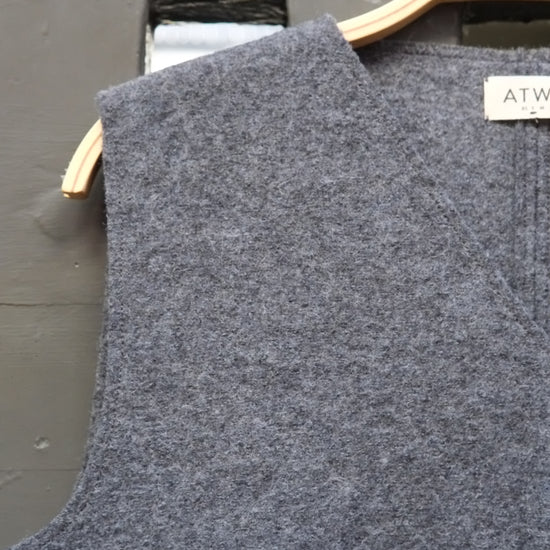 A close-up of a grey sleeveless knit top hanging on a light wood hanger against a dark grey wall. A small white brand label reading Atwin is visible near the neckline.