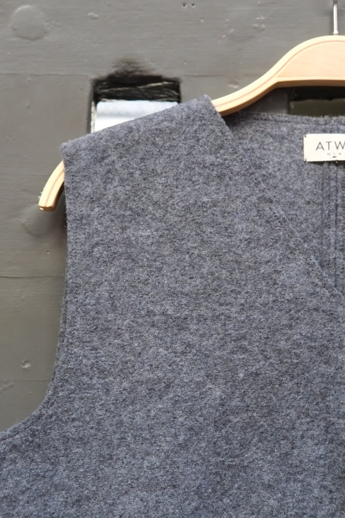 A close-up of a grey sleeveless knit top hanging on a light wood hanger against a dark grey wall. A small white brand label reading Atwin is visible near the neckline.