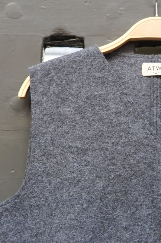 A close-up of a grey sleeveless knit top hanging on a light wood hanger against a dark grey wall. A small white brand label reading Atwin is visible near the neckline.
