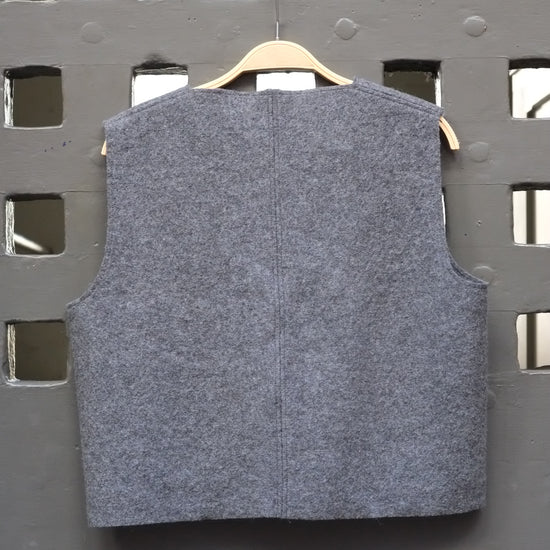 A sleeveless grey boiled wool vest hanging on a wooden hanger against a dark grey metal lattice door.