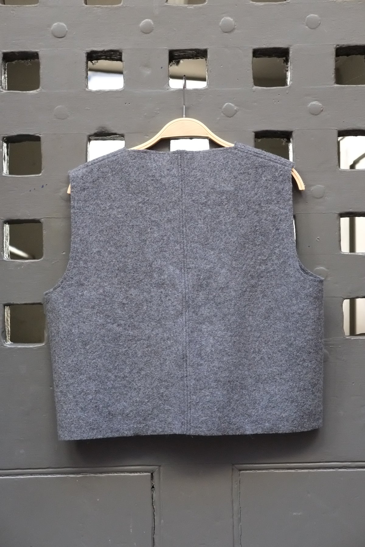 A sleeveless grey boiled wool vest hanging on a wooden hanger against a dark grey metal lattice door.