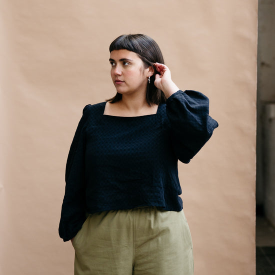 Model with dark short hair wearing a black top and khaki trousers against a beige background