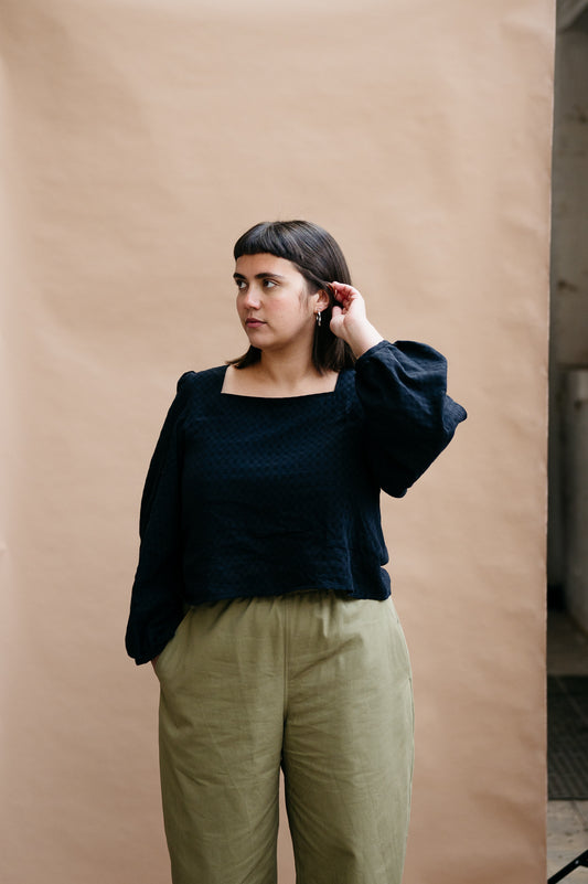 Model with dark short hair wearing a black top and khaki trousers against a beige background