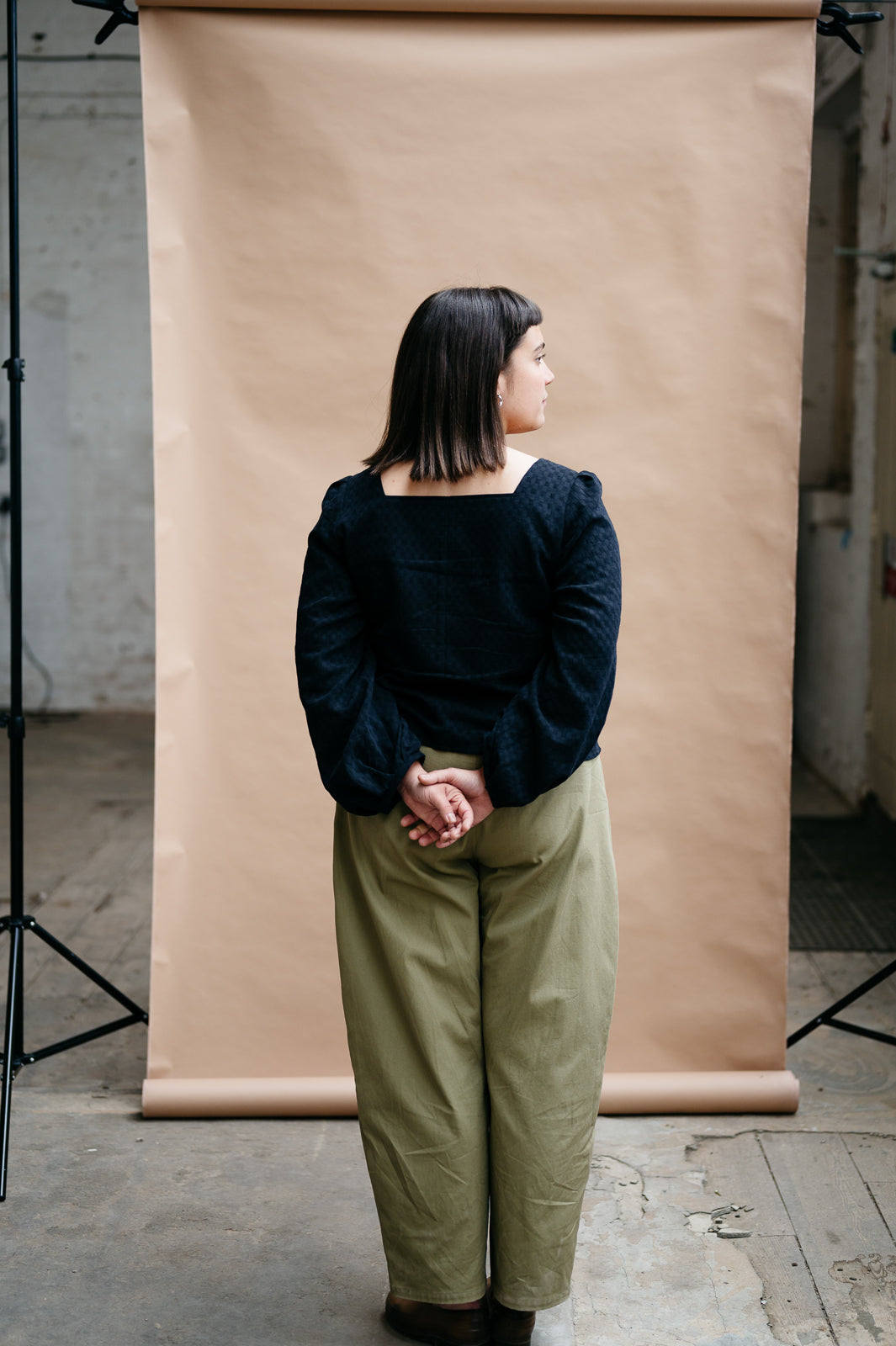 Person wearing a black square neck long sleeve top and khaki trousers standing in front of a beige backdrop