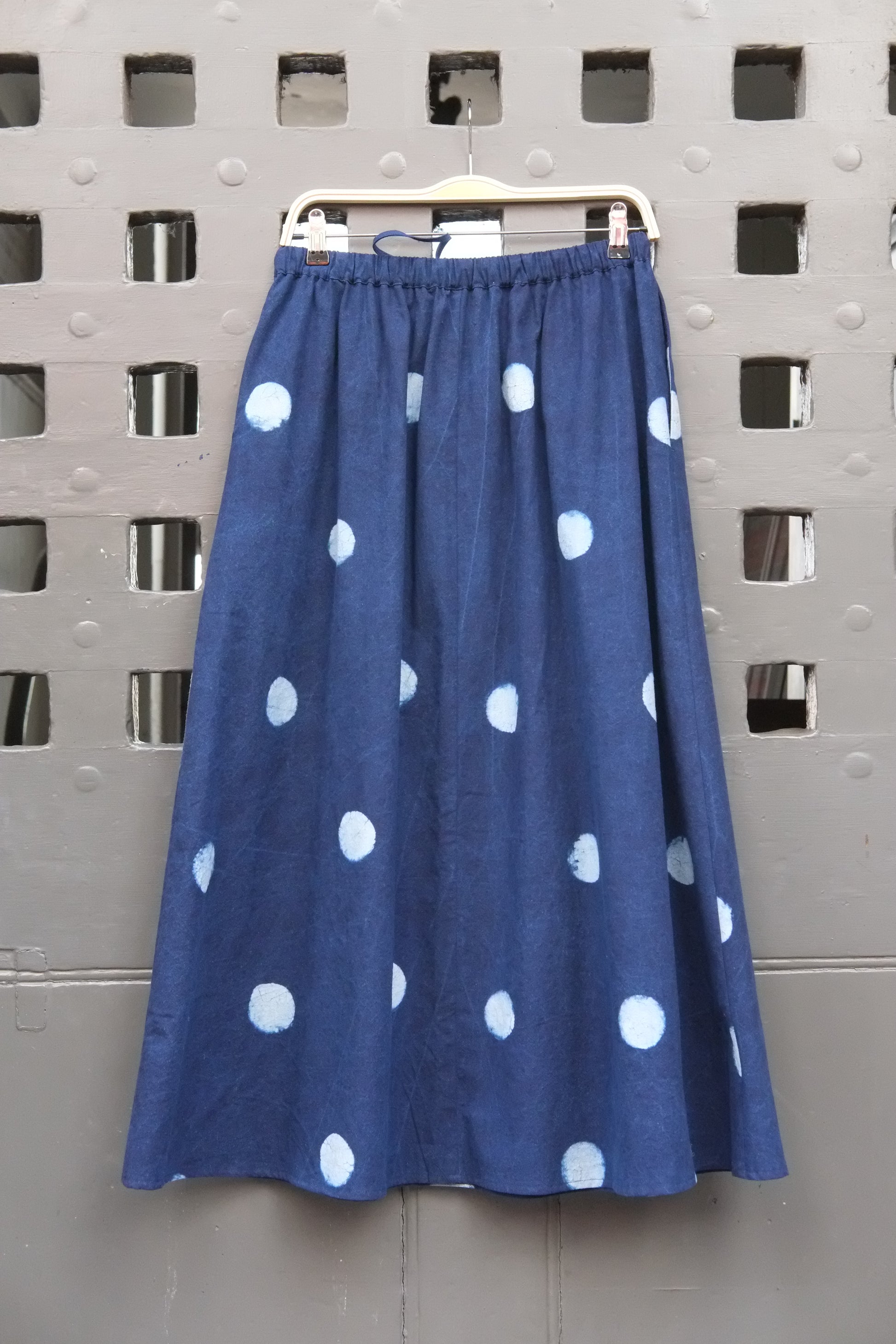 Polka dot indigo and pale blue block printed long skirt with drawstring waist tie on a wooden hanger in front of a grey grid door