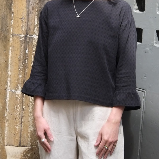 Person wearing a black textured blouse with white trousers against a stone wall.