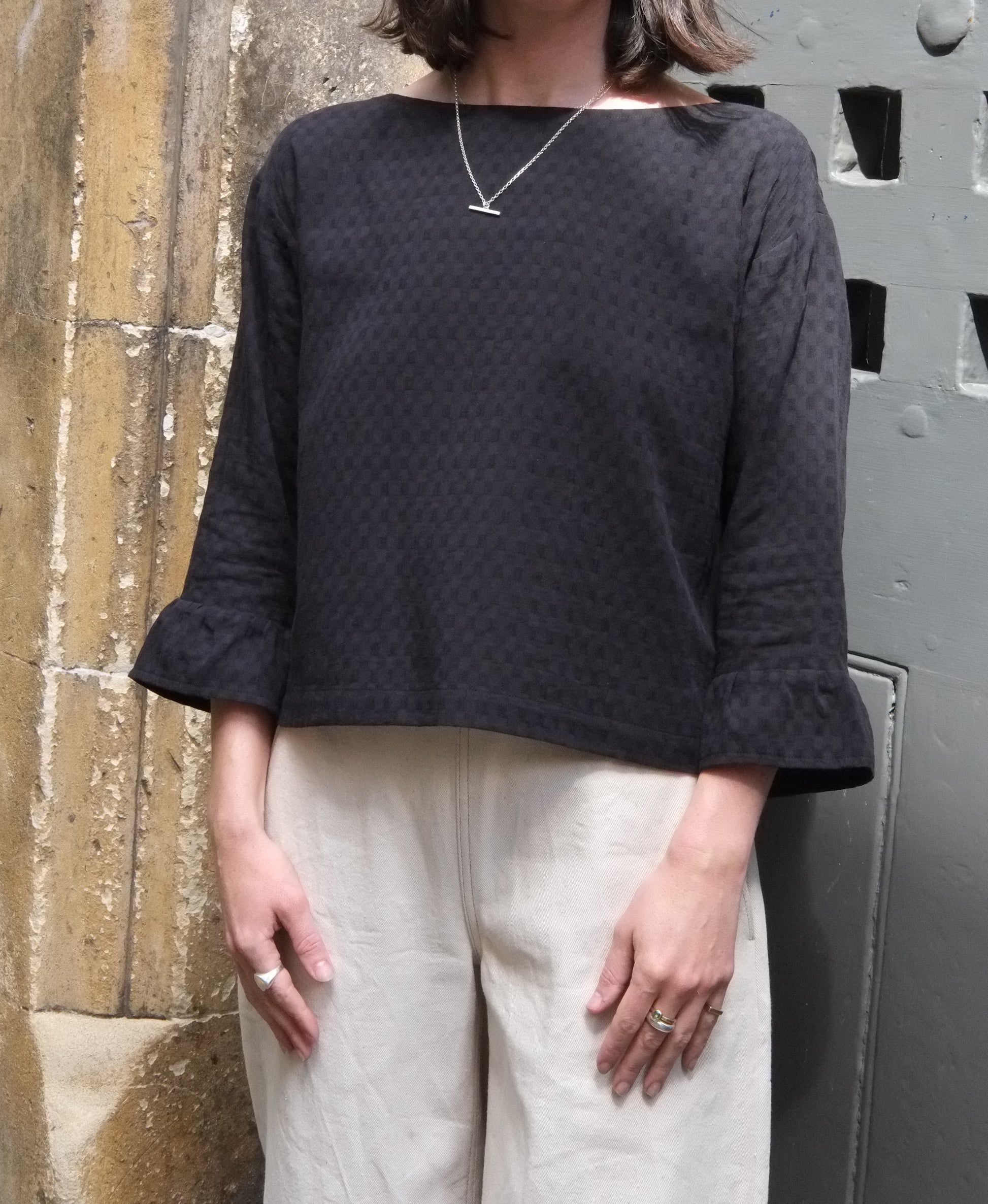 Person wearing a black textured blouse with white trousers against a stone wall.