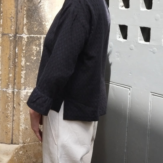 Person wearing a black frill sleeve top and light trousers against a textured wall.