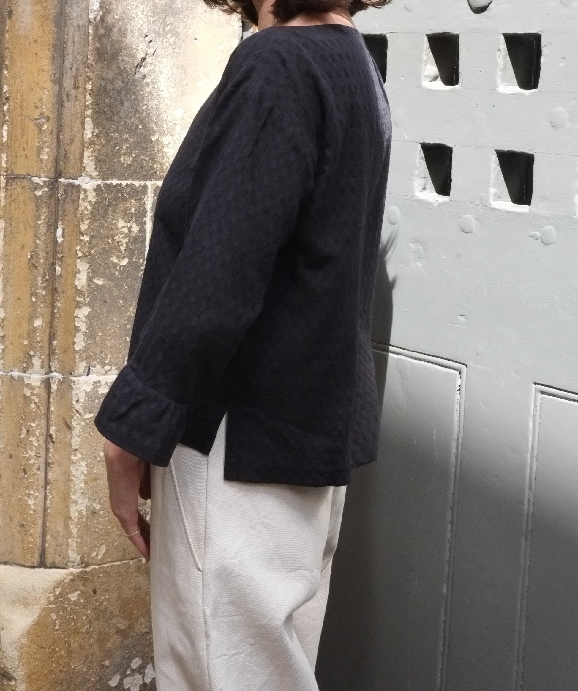 Person wearing a black frill sleeve top and light trousers against a textured wall.