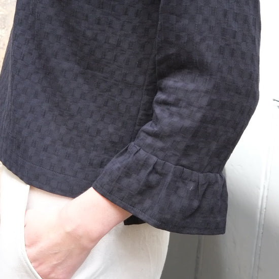 Close-up of a person wearing black patterned top with white trousers against a grey background