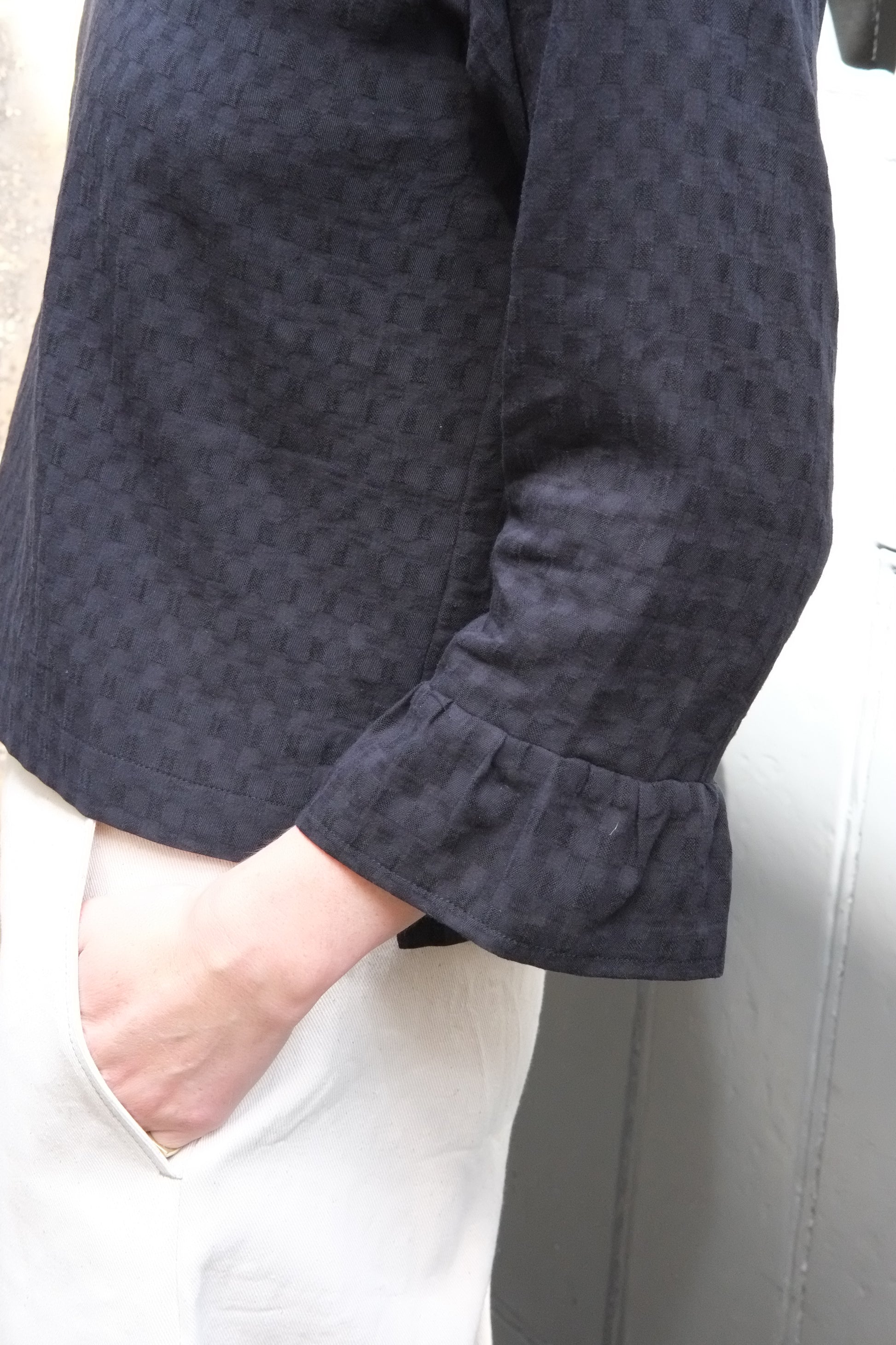 Close-up of a person wearing black patterned top with white trousers against a grey background