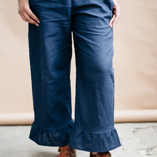 Person wearing navy blue trousers with ruffled cuffs against a beige background and brown sandals