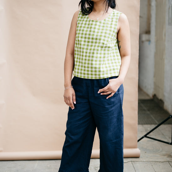 Woman wearing a green checkered top and navy blue trousers with a frill hem standing against a beige wall.