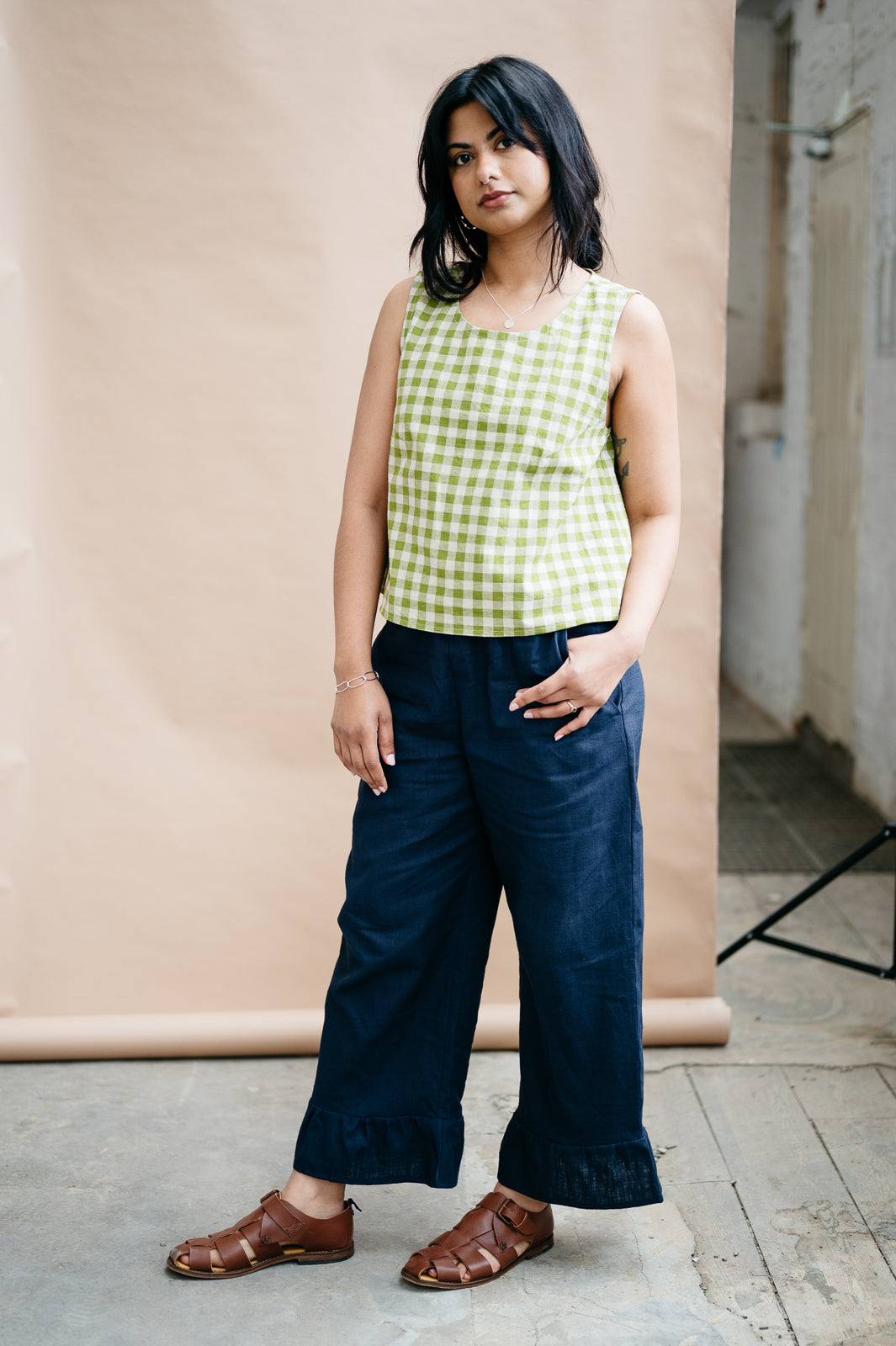 Woman wearing a green checkered top and navy blue trousers with a frill hem standing against a beige wall.