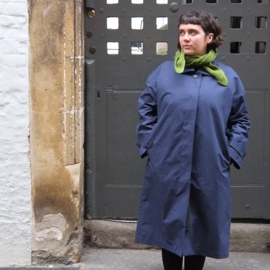 Person wearing a blue coat and green scarf standing on a sidewalk.